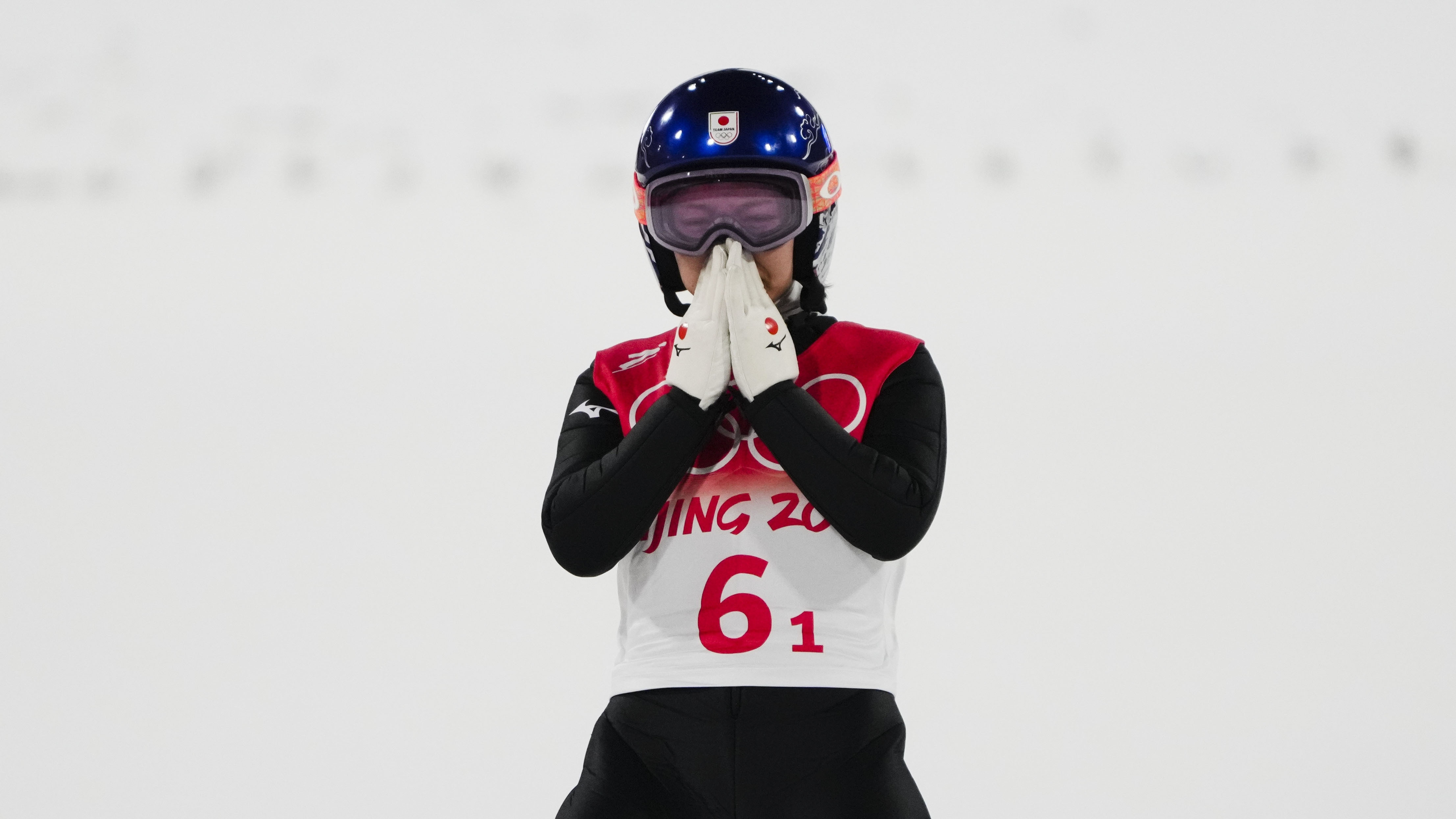 A woman wearing Olympic ski gear, including a helmet, holds her hands in front of her nose and mouth.